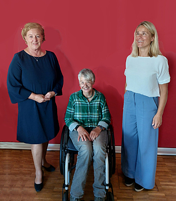 Foto: Michaela Engelmeier in dunkelblauem knielangem Kleid, Sigrid Arnade in grünem kariertem Hemd und Jeans sitzt im Rollstuhl, Verena Bentele in hellblauer Schlaghose und weißem kurzärmeligem Top. Im Hintergrund eine tiefrote Wand