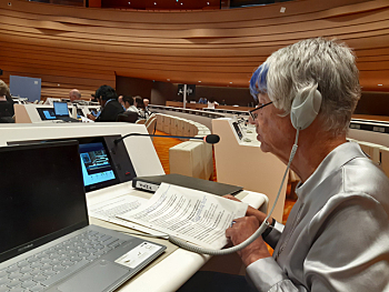 Foto Sigrid Arnade, sie sitzt an einem Delegiertenplatz im Plenarsaal und spricht mit Blick in ihre Unterlagen in ein Platzmikrofon und hat einen großen grauen Ohrhörer auf.