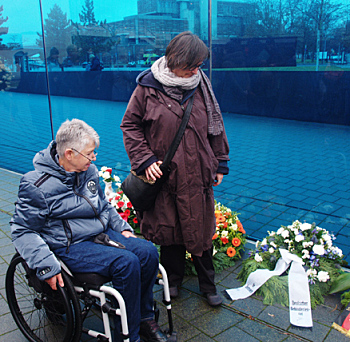 Foto: Sigrid Arnade (mit Rollstuhl) und Martina Puschke (stehend) blicken auf den Kranz des Deutschen Behindertenrates an der Blauen Wand der T4-Gedenkstätte