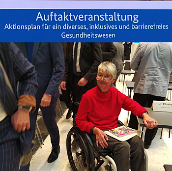 Sigrid Arnade sitz im Rollstuhl im Tagungsraum. Sie hat Informationsmaterial auf den Knien und lächelt skeptisch in die Kamera. An ihr vorbei gehen Menschen in Herrenanzügen, durch den Bildausschnitt sind keine Gesichter zu sehen.