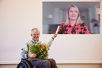 Staffelstabübergabe - Prof. Dr. Sigrid Arnade, Weibernetz, sitzt im Rollstuhl mit einem Blumenstrauß auf dem Schoß und übergibt den Staffelstab an Verena Bentele, Sozialverband VdK Deutschland, diese ist per Video eingeblendet